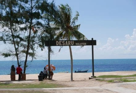 Visitors rest beneath a Desaru beach sign beside a sandy shore and the sea.